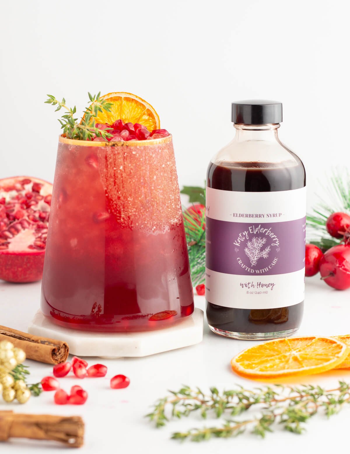 A glass of red punch garnished with orange slice, pomegranate seeds, and herbs sits beside a bottle of elderberry syrup. Pomegranate, cinnamon sticks, dried citrus, and festive decorations are in the background.