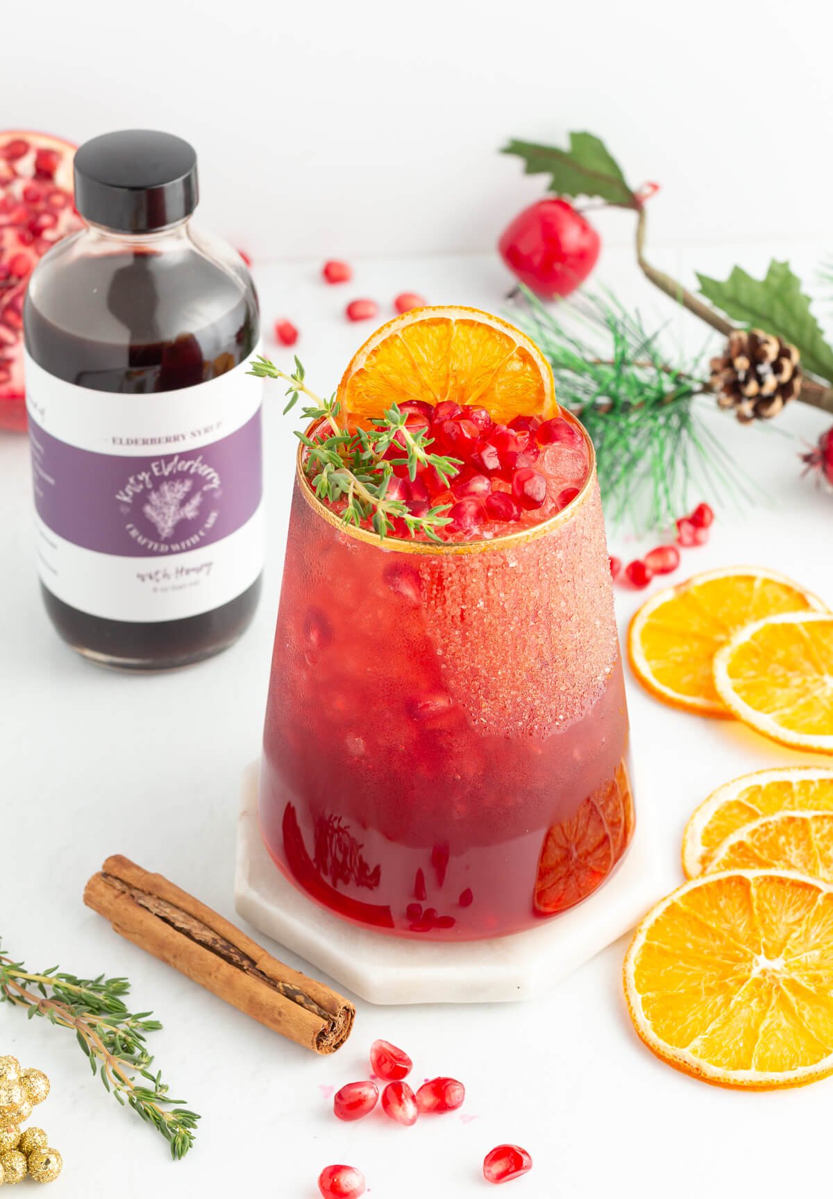 A glass of red iced drink garnished with orange slices, pomegranate seeds, and a sprig of thyme sits on a white coaster. Nearby are dried orange slices, cinnamon stick, a bottle of elderberry syrup, and festive greenery.