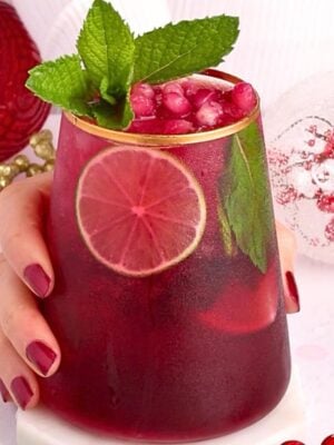 A hand with red polished nails holds a glass of red drink garnished with a lime slice, pomegranate seeds, and mint leaves; holiday ornaments are in the background.