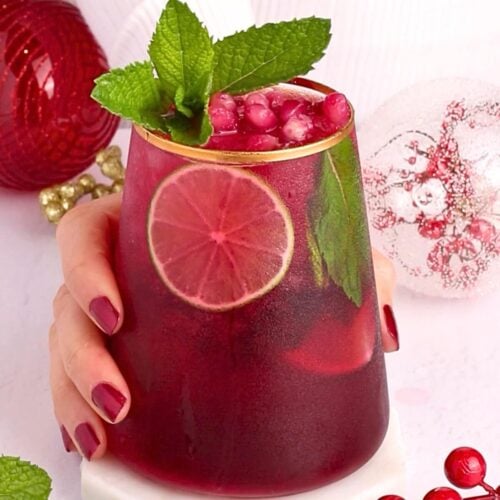 A hand with red polished nails holds a glass of red drink garnished with a lime slice, pomegranate seeds, and mint leaves; holiday ornaments are in the background.
