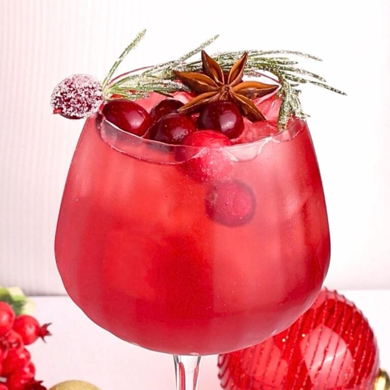 A festive red cocktail in a large glass, garnished with cranberries, a sugared cranberry, a sprig of rosemary, and a star anise. The drink is surrounded by holiday decorations.