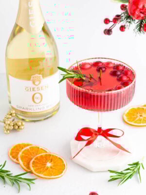 A festive red cocktail garnished with cranberries and rosemary in a glass tied with a red ribbon, surrounded by dried orange slices, rosemary sprigs, and a bottle of Giesen sparkling brut. Holiday decorations are in the background.