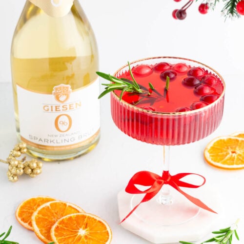 A festive red cocktail garnished with cranberries and rosemary in a glass tied with a red ribbon, surrounded by dried orange slices, rosemary sprigs, and a bottle of Giesen sparkling brut. Holiday decorations are in the background.
