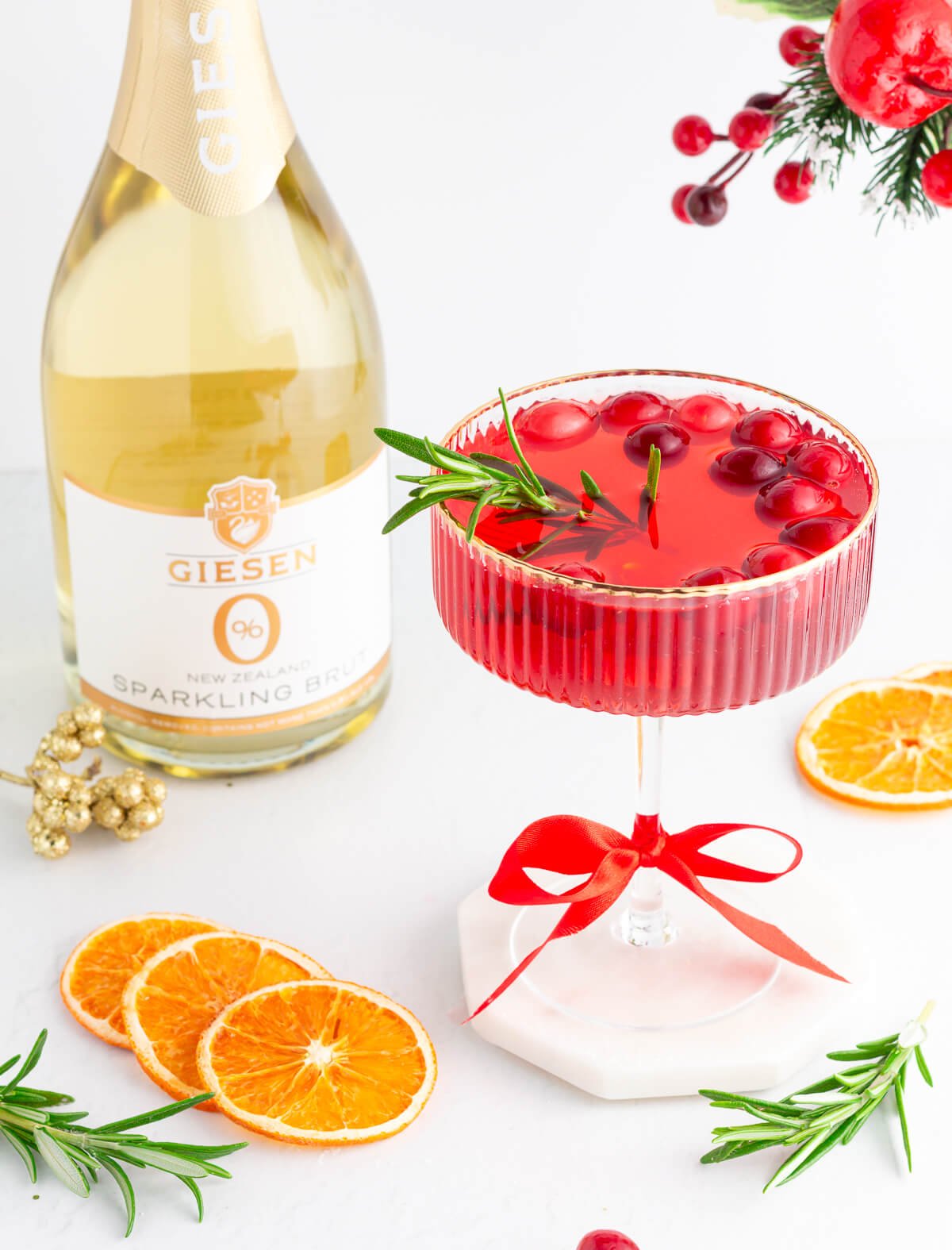 A festive red cocktail garnished with cranberries and rosemary in a glass tied with a red ribbon, surrounded by dried orange slices, rosemary sprigs, and a bottle of Giesen sparkling brut. Holiday decorations are in the background.