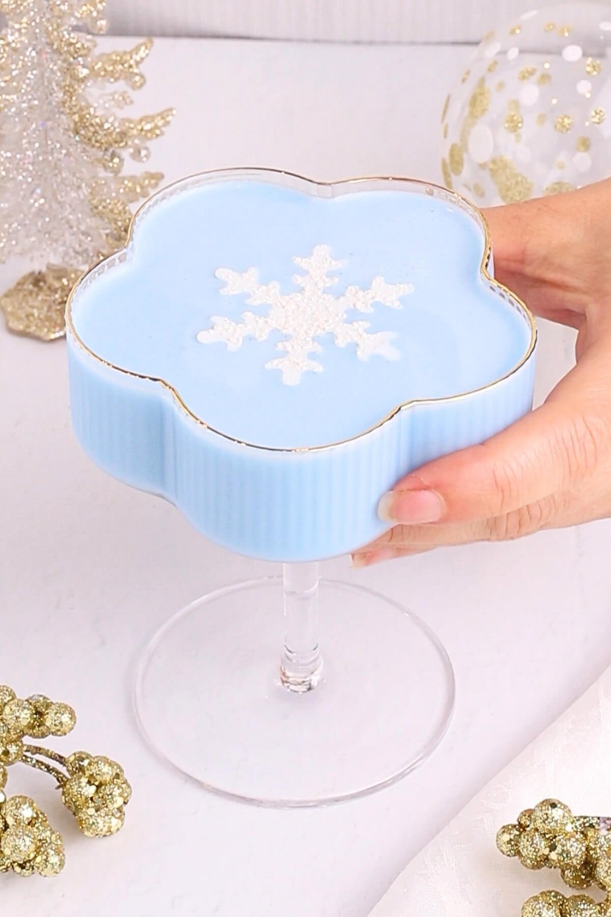 A hand holds a blue, flower-shaped dessert in a glass cup, topped with a white snowflake decoration. The background features festive gold and white holiday decor.