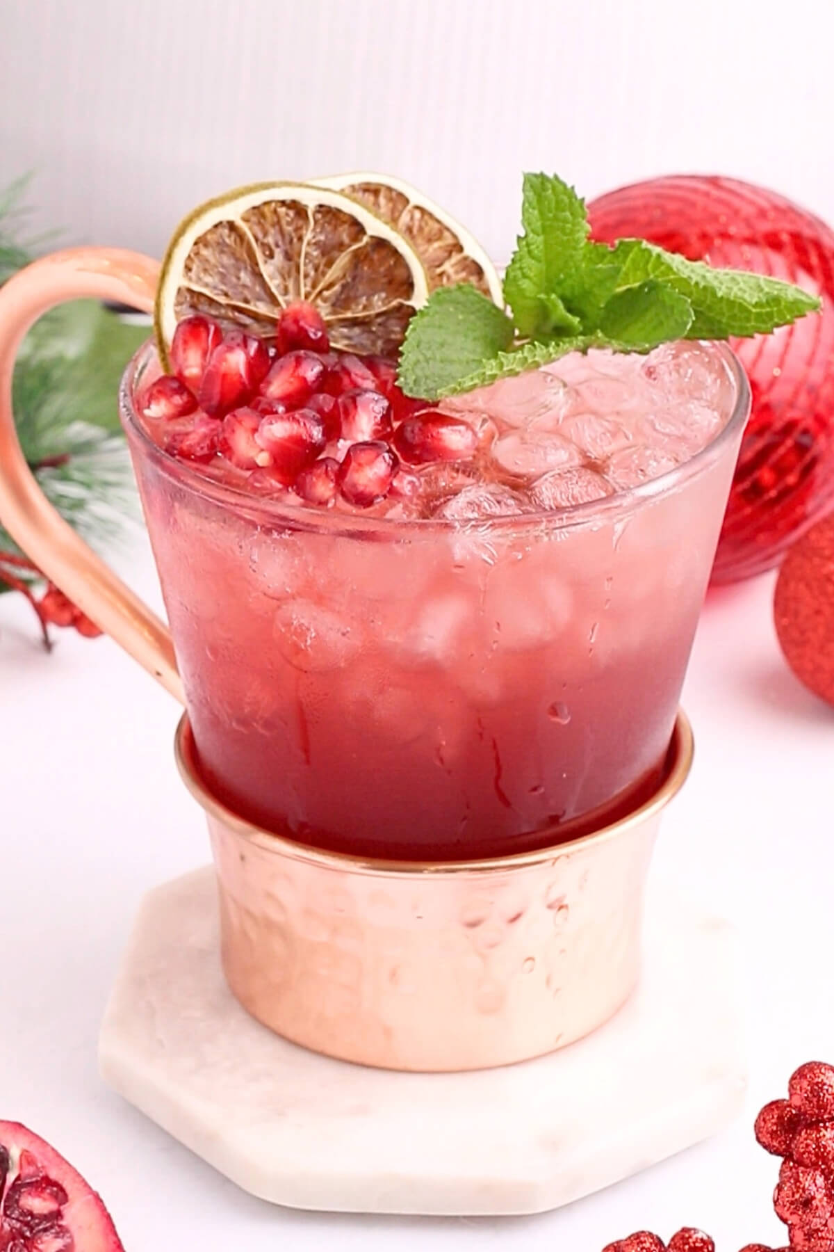 A glass mug filled with a red iced drink, garnished with pomegranate seeds, lime slices, and a sprig of mint, sits on a marble coaster. Red ornaments and pomegranate pieces are visible in the background.