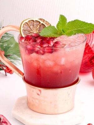 A festive drink in a copper mug garnished with mint, a lime slice, and pomegranate seeds, surrounded by holiday decorations, a pine branch, and a pomegranate half on a white surface.