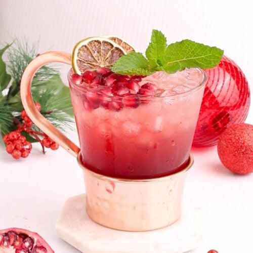 A festive drink in a copper mug garnished with mint, a lime slice, and pomegranate seeds, surrounded by holiday decorations, a pine branch, and a pomegranate half on a white surface.