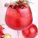 A large glass of red spritz mocktail with ice, cranberries, and garnishes of sugared rosemary, star anise, and a sugared cranberry. Decorative ornaments and berries are arranged around the base of the glass.