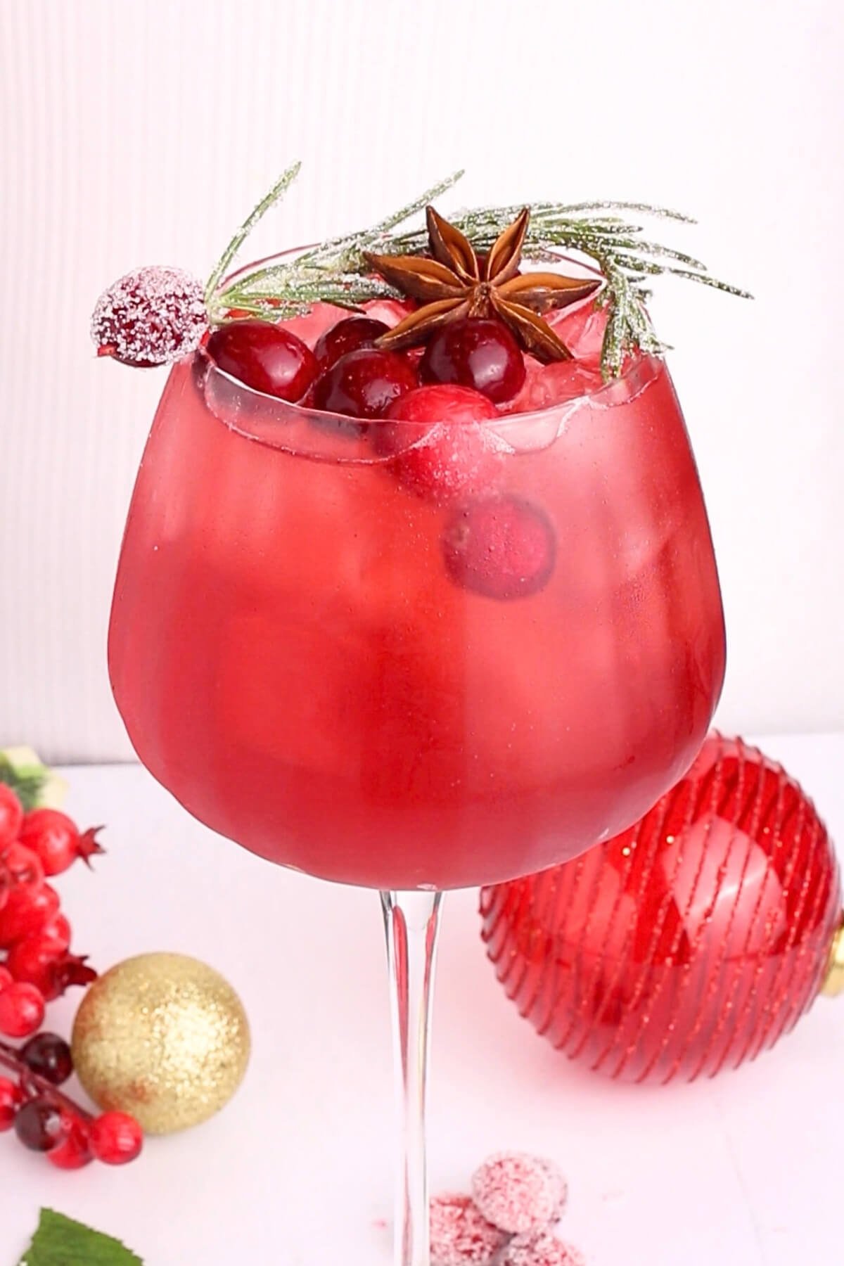 A large glass of red spritz mocktail with ice, cranberries, and garnishes of sugared rosemary, star anise, and a sugared cranberry. Decorative ornaments and berries are arranged around the base of the glass.