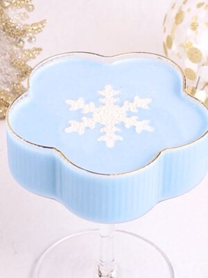 A light blue mocktail in a flower-shaped glass, topped with a white snowflake design. The background features white and gold holiday decorations, including a tree and an ornament.