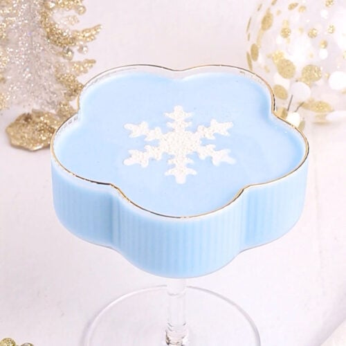 A light blue mocktail in a flower-shaped glass, topped with a white snowflake design. The background features white and gold holiday decorations, including a tree and an ornament.