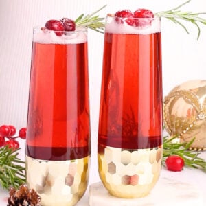 Two tall glasses of red sparkling drink garnished with cranberries and rosemary sit on a white surface, surrounded by cranberries, a pinecone, greenery, and a gold ornament. The glasses have gold geometric bases.