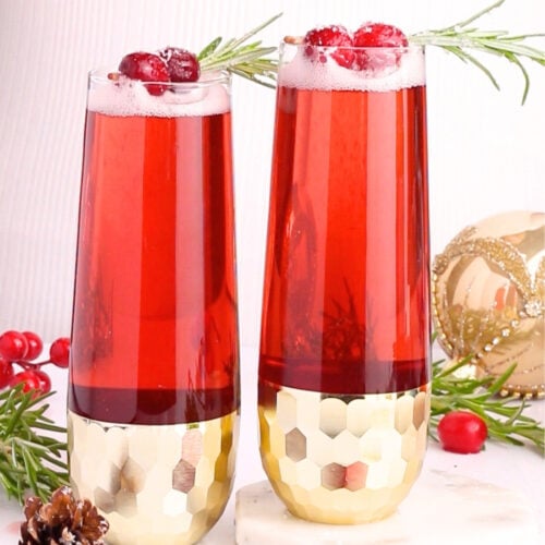 Two tall glasses of red sparkling drink garnished with cranberries and rosemary sit on a white surface, surrounded by cranberries, a pinecone, greenery, and a gold ornament. The glasses have gold geometric bases.