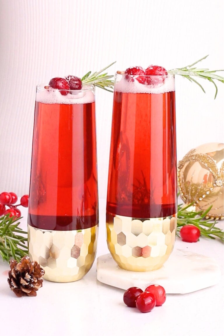 Two tall glasses of red sparkling drink garnished with cranberries and rosemary sit on a white surface, surrounded by cranberries, a pinecone, greenery, and a gold ornament. The glasses have gold geometric bases.