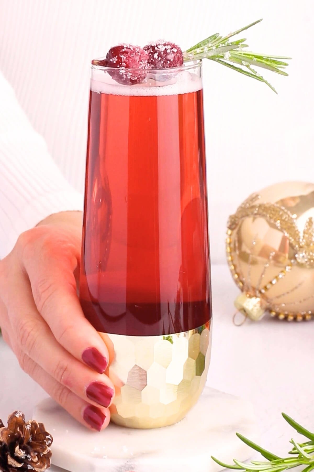 A hand with red-painted nails holds a tall glass of red holiday drink, garnished with sugared cranberries and a sprig of rosemary. A gold ornament and pinecone are in the background.