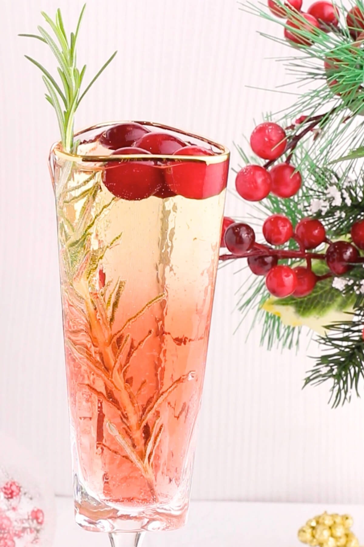 A tall glass filled with a pink fizzy drink, garnished with cranberries and a sprig of rosemary. Festive greenery and red berry decorations are visible in the background.