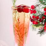 A glass of sparkling drink garnished with rosemary and cranberries, with festive red berries and pine branches in the background.