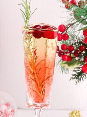 A glass of sparkling drink garnished with rosemary and cranberries, with festive red berries and pine branches in the background.