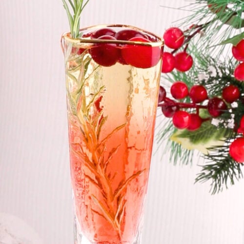 A glass of sparkling drink garnished with rosemary and cranberries, with festive red berries and pine branches in the background.