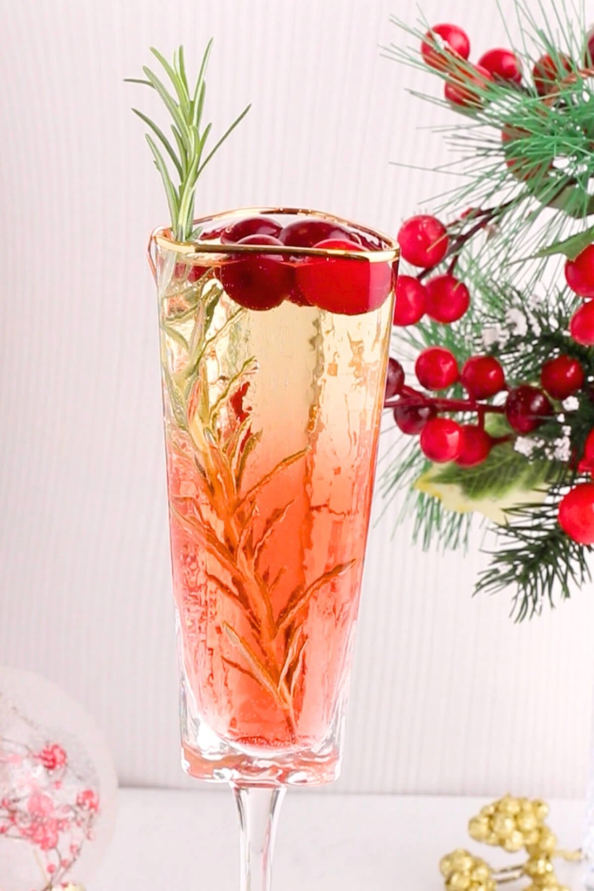 A glass of sparkling drink garnished with rosemary and cranberries, with festive red berries and pine branches in the background.