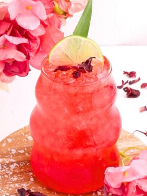 A vibrant red drink with crushed ice is served in a curvy glass, garnished with a lime slice, hibiscus petals, and surrounded by pink flowers on a wooden surface.