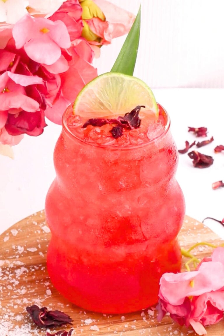 A vibrant red drink with crushed ice is served in a curvy glass, garnished with a lime slice, hibiscus petals, and surrounded by pink flowers on a wooden surface.