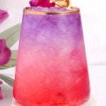 A vibrant mocktail with a gradient from pink at the bottom to purple at the top, served in a glass with gold rim, garnished with a dried citrus slice and a small purple flower, sitting on a white coaster.