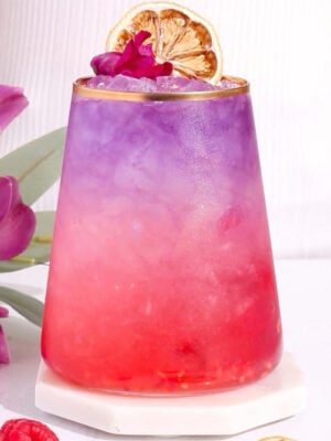 A vibrant mocktail with a gradient from pink at the bottom to purple at the top, served in a glass with gold rim, garnished with a dried citrus slice and a small purple flower, sitting on a white coaster.