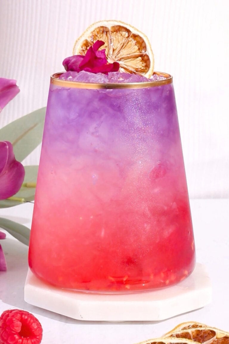 A vibrant mocktail with a gradient from pink at the bottom to purple at the top, served in a glass with gold rim, garnished with a dried citrus slice and a small purple flower, sitting on a white coaster.