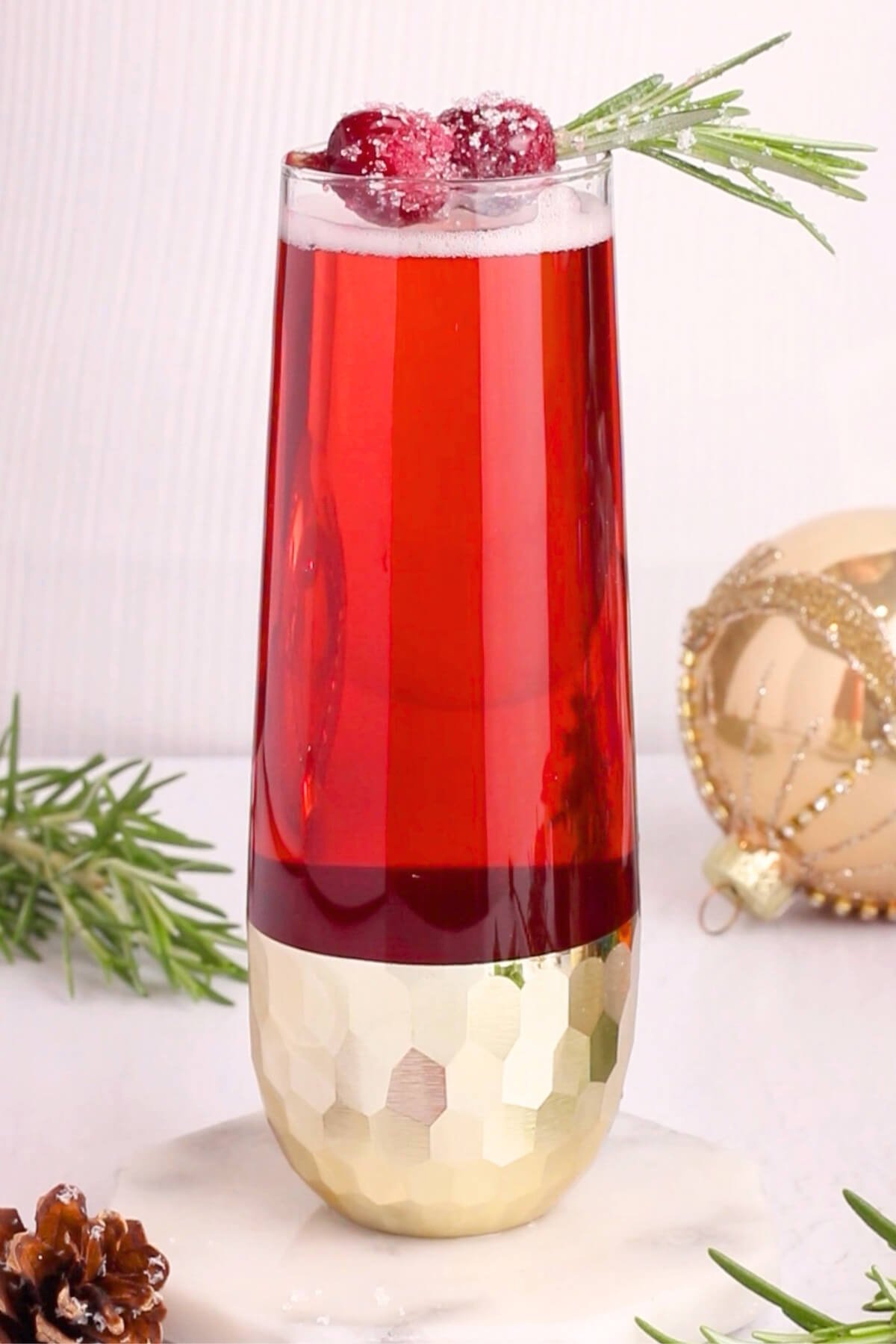 A tall glass of red drink garnished with sugared cranberries and a rosemary sprig sits on a white surface. The glass has a gold textured base. Holiday decorations, including a gold ornament, are in the background.
