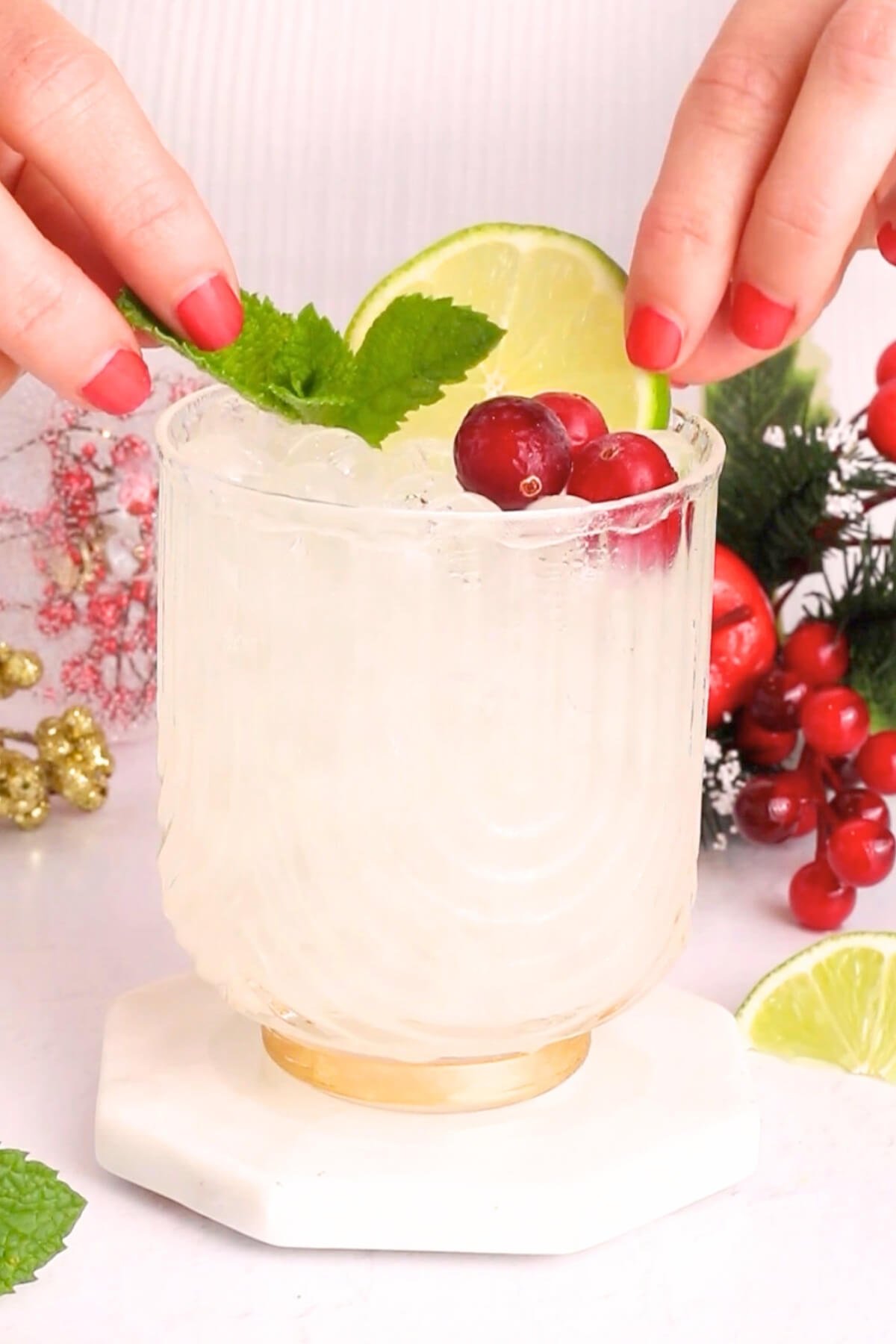 A hand with red nail polish garnishes a clear, textured glass of ice-filled drink with a mint sprig, cranberries, and a lime slice; festive decorations and lime wedges are in the background.