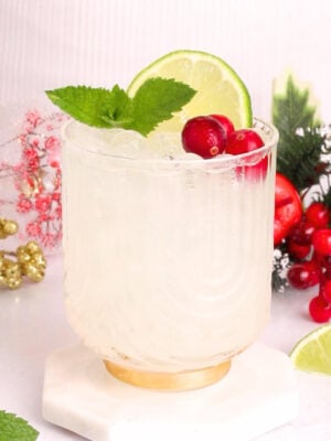 A clear glass filled with a white mocktail is garnished with a lime slice, fresh mint leaves, and red cranberries. In the background, there are festive holiday decorations and more cranberries and lime slices.