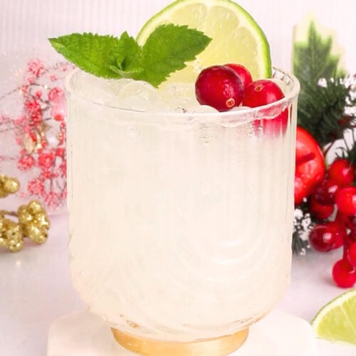 A clear glass filled with a white mocktail is garnished with a lime slice, fresh mint leaves, and red cranberries. In the background, there are festive holiday decorations and more cranberries and lime slices.