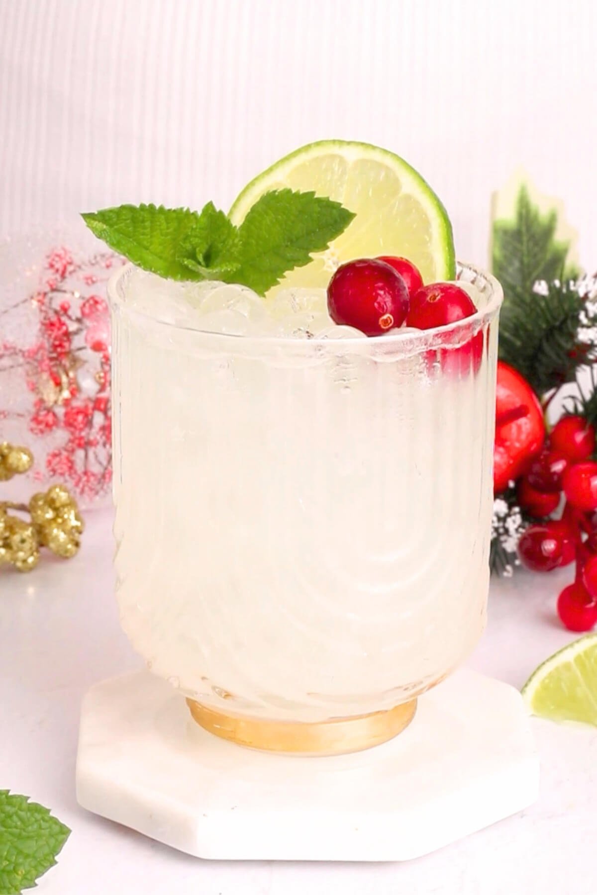 A clear glass filled with a white mocktail is garnished with a lime slice, fresh mint leaves, and red cranberries. In the background, there are festive holiday decorations and more cranberries and lime slices.