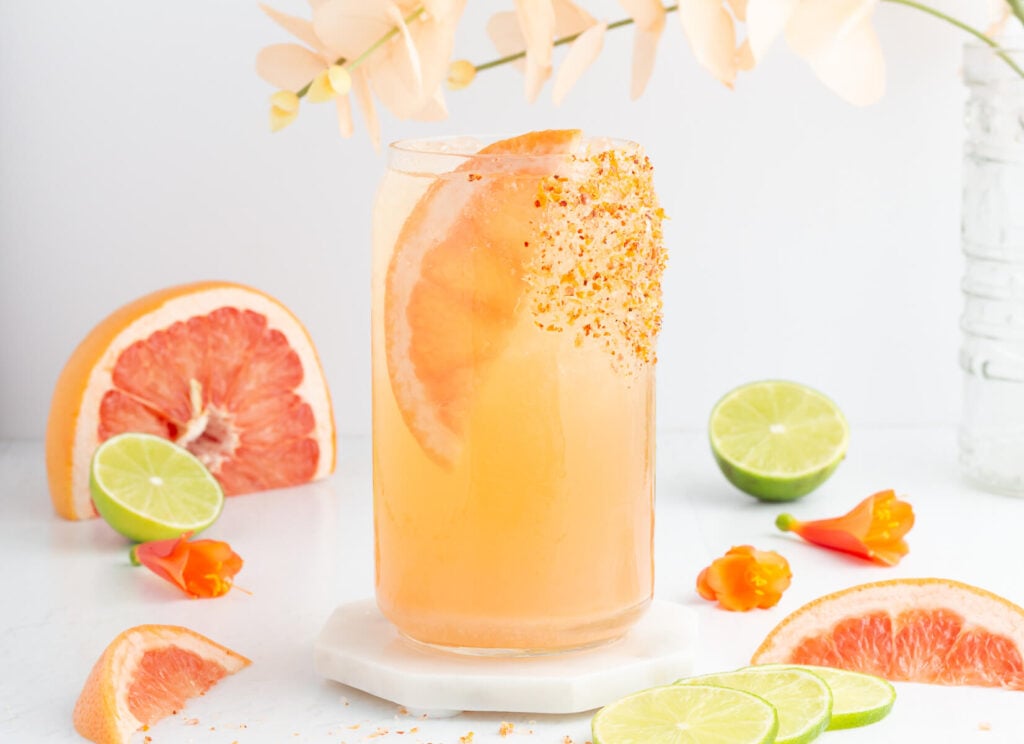A tall glass of grapefruit drink garnished with a grapefruit slice and chili powder on the rim, surrounded by lime halves, grapefruit wedges, orange peppers, and pale flowers on a white surface.