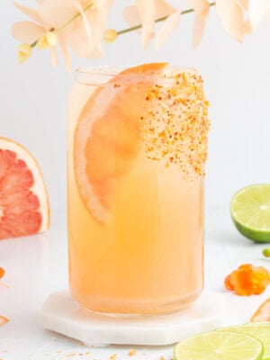 A tall glass of grapefruit drink garnished with a grapefruit slice and chili powder on the rim, surrounded by lime halves, grapefruit wedges, orange peppers, and pale flowers on a white surface.