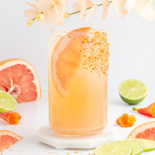 A tall glass of grapefruit drink garnished with a grapefruit slice and chili powder on the rim, surrounded by lime halves, grapefruit wedges, orange peppers, and pale flowers on a white surface.