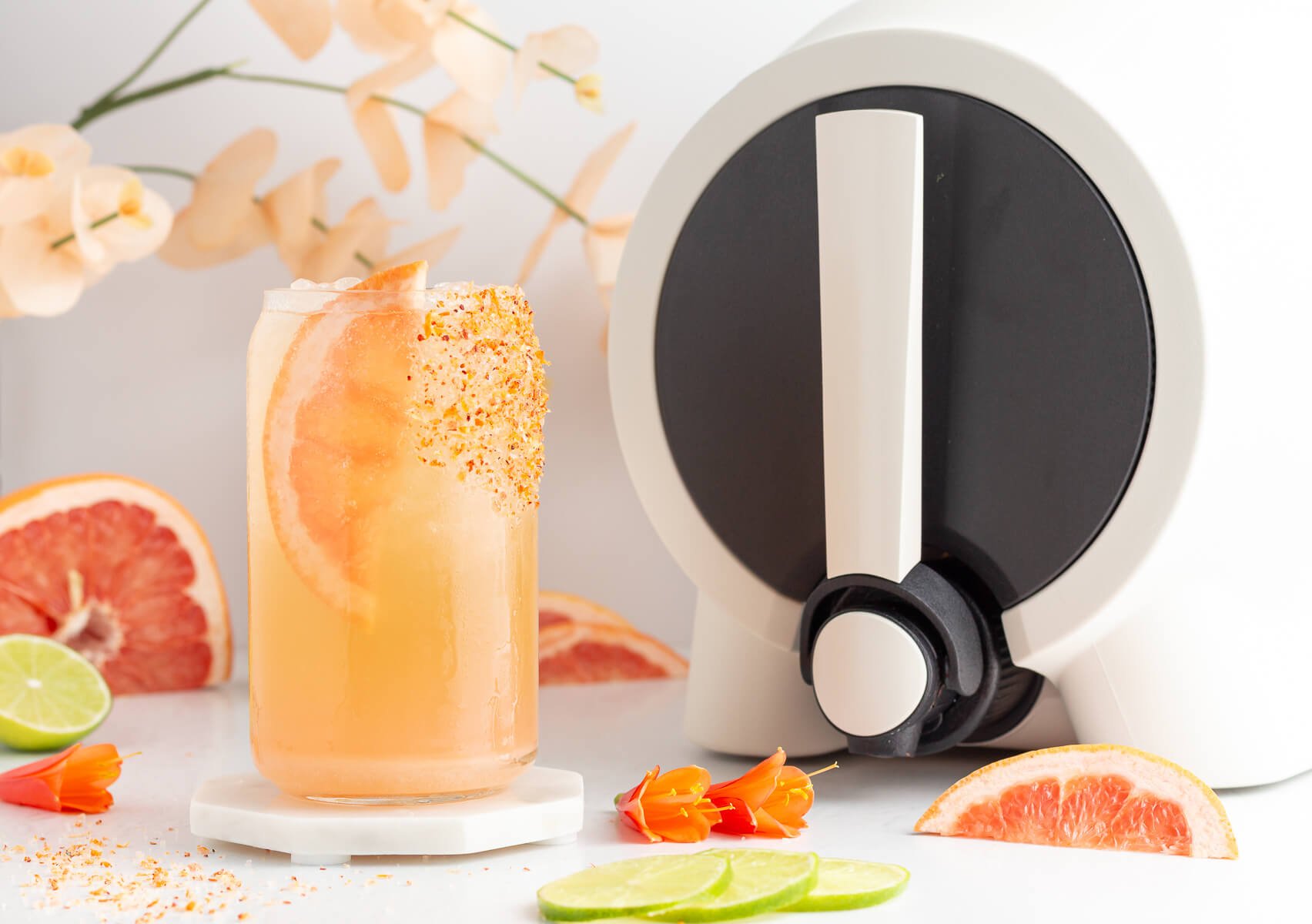 A tall glass of grapefruit mocktail with a chili-salt rim and a grapefruit slice, surrounded by lime slices, grapefruit wedges, orange flowers, and a modern white drink dispenser in the background.