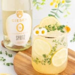 A glass of sparkling lemon and herb spritz garnished with lemon slices, thyme, and small white flowers sits on a wooden board next to a bottle of Giesen 0% Sauvignon Blanc Spritz. Bright greenery decorates the background.