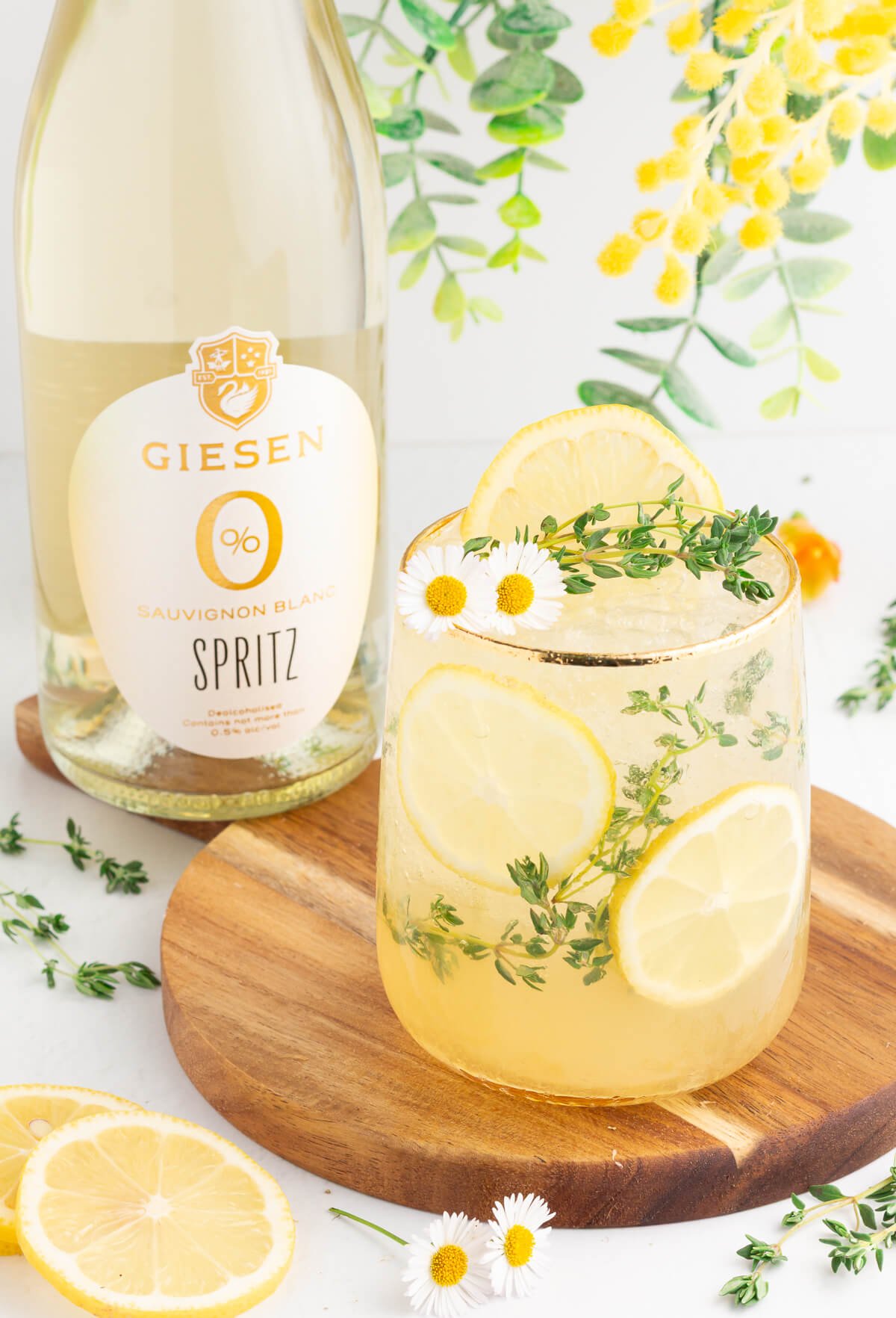 A glass of sparkling lemon and herb spritz garnished with lemon slices, thyme, and small white flowers sits on a wooden board next to a bottle of Giesen 0% Sauvignon Blanc Spritz. Bright greenery decorates the background.