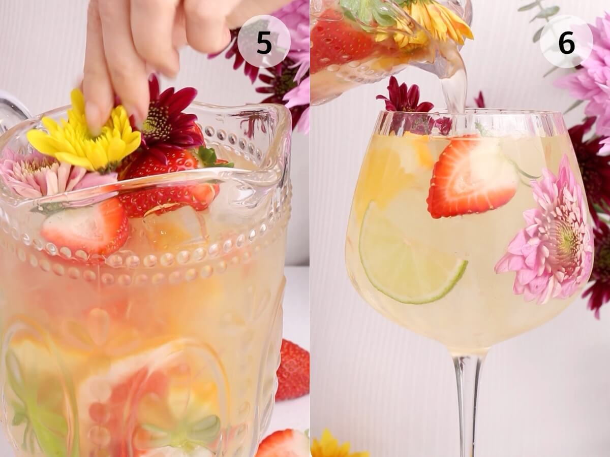 A hand adds colorful flowers and fruit slices to a clear pitcher of sangria. Next to it, sangria with fruit and flowers is poured into a large wine glass. Pink flowers and strawberries are visible in the scene.