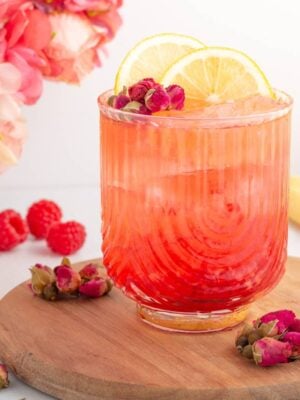A vibrant pink drink with ice in a ribbed glass, garnished with lemon slices and small rosebuds, sits on a wooden board. Fresh raspberries, rosebuds, and a sliced lemon surround the glass, with flowers in the background.