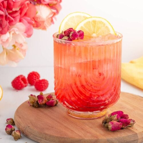 A vibrant pink drink with ice in a ribbed glass, garnished with lemon slices and small rosebuds, sits on a wooden board. Fresh raspberries, rosebuds, and a sliced lemon surround the glass, with flowers in the background.