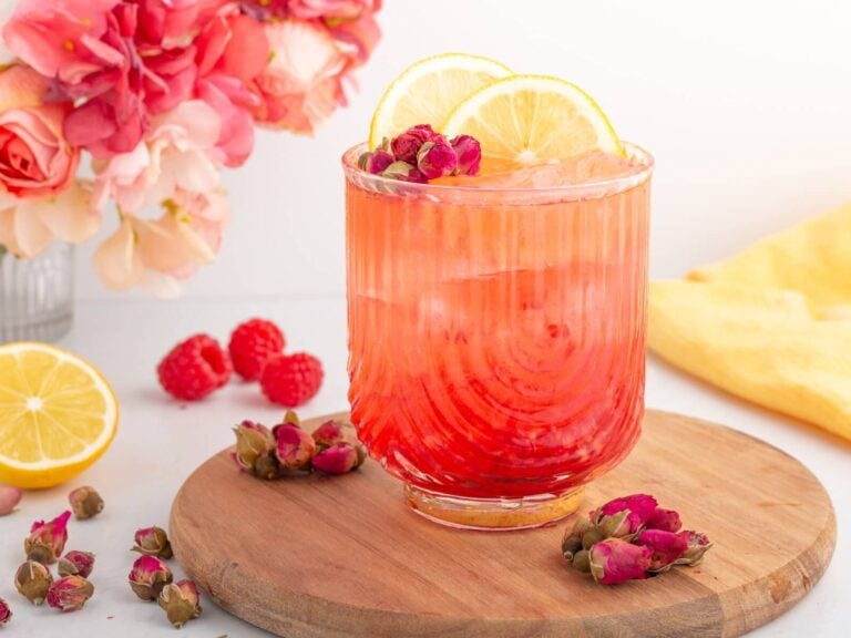A vibrant pink drink with ice in a ribbed glass, garnished with lemon slices and small rosebuds, sits on a wooden board. Fresh raspberries, rosebuds, and a sliced lemon surround the glass, with flowers in the background.