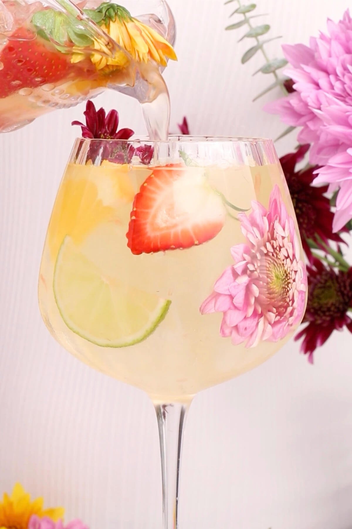A glass of clear, refreshing drink with floating lime and strawberry slices and pink flower petals, surrounded by colorful flowers, as liquid is being poured in.