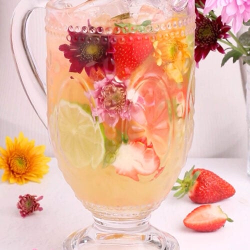 A glass pitcher filled with a light yellow drink, ice cubes, sliced limes, strawberries, and pink flowers. Fresh flowers, strawberries, and a lime wedge are arranged around the pitcher on a white surface.