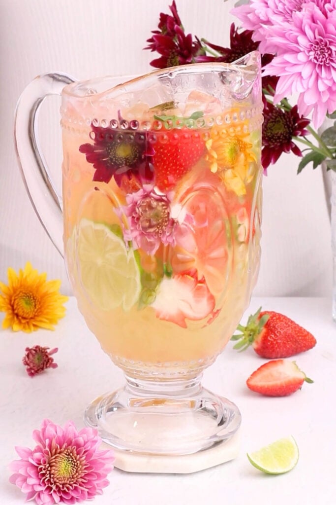 A glass pitcher filled with a light yellow drink, ice cubes, sliced limes, strawberries, and pink flowers. Fresh flowers, strawberries, and a lime wedge are arranged around the pitcher on a white surface.