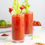 Two tall glasses of Bloody Mary cocktails garnished with celery stalks, lime wedges, and cherry tomatoes sit on a white surface with limes, tomatoes, and a small bowl of red spice in the background.
