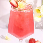 A pink mocktail with ice in a stemmed glass, garnished with a lemon slice, half a strawberry, and a yellow flower. Fresh strawberries and dried lemon slices are on the white table nearby.